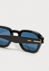 Μαύρα γυαλιά ηλίου με μπλε φακούς, τετράγωνο σκελετό και χρυσές λεπτομέρειες στους ναούς. Χαρακτηρίζονται από τολμηρό, μοντέρνο σχέδιο.