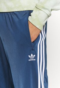 Námořnické sportovní kalhoty mají zipovou kapsu, bílé pruhy po stranách a bílé logo Adidas v levém dolním rohu. Hladká, lesklá tkanina.