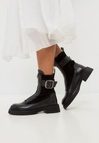 Zwarte enkelboots met een mix van glad en textuurleer, ritsluiting, zijgesp voor extra detail, en een chunky, geribbelde zool.