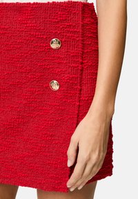 Red textured mini skirt with a woven appearance, featuring two gold buttons on the front and a side slit for added detail.