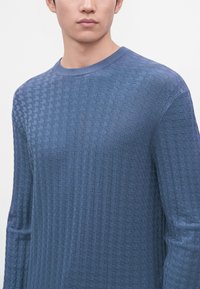 Jeune homme portant un pull bleu texturé à manches longues et col rond, montré de la poitrine au menton sur un fond uni.