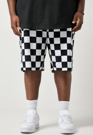 Person iført sort-hvide ternede shorts, hvide sokker og hvide sneakers, stående mod en ensfarvet lys baggrund.
