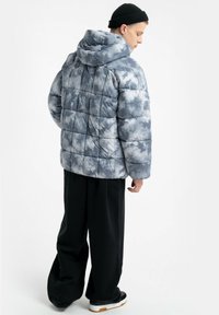 Kurtka puchowa z kapturem o pikowanym wzorze w szarym stylu tie-dye. Miękka tekstura, luźny krój, z ściągaczami przy mankietach, noszona z luźnymi czarnymi spodniami i trampkami.