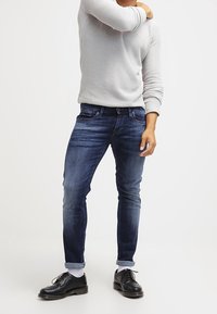 Mörkblå denimjeans med slim fit, med uppvikta benslut. Bärs med en ljusgrå stickad tröja och svarta snörskor.