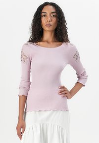 Lila geribbelde trui met 3/4 mouwen, golvende zoom en bloemige kantaccenten op de schouders, gecombineerd met een witte rok.