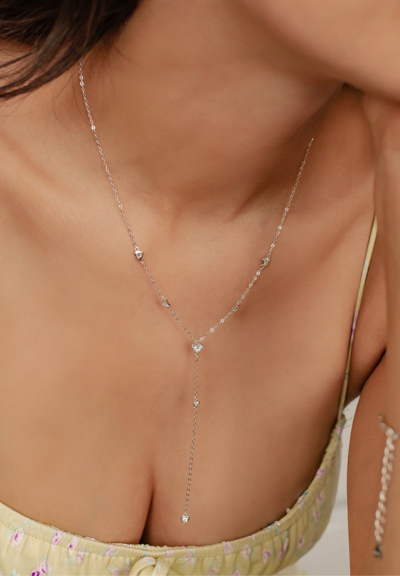 Collana in argento con accenti a forma di cuore, caratterizzata da un design a Y e delicate maglie, indossata attorno al collo su uno sfondo neutro.