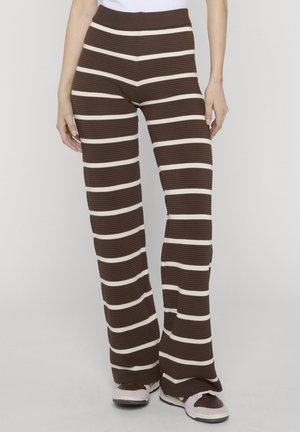 Person wearing brown wide-leg pants with white horizontal stripes and white sneakers, standing against a plain background.