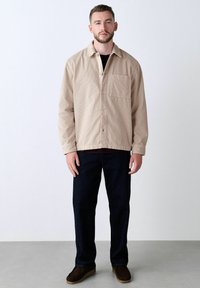 Man standing wearing beige corduroy jacket, dark pants, and brown shoes against plain white background.