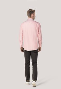 Gestreiftes rosa Langarmshirt mit Button-Down-Kragen, kombiniert mit dunkelgrauen Hosen und weißen Turnschuhen. Einfarbiger Hintergrund.