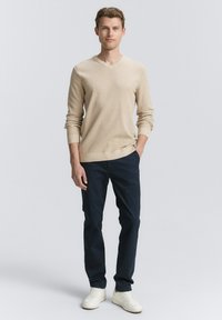 Beiger V-Ausschnitt-Pullover aus strukturiertem Stoff, kombiniert mit marineblauen Hosen und weißen Sneakers, die eine schlanke Silhouette präsentieren.