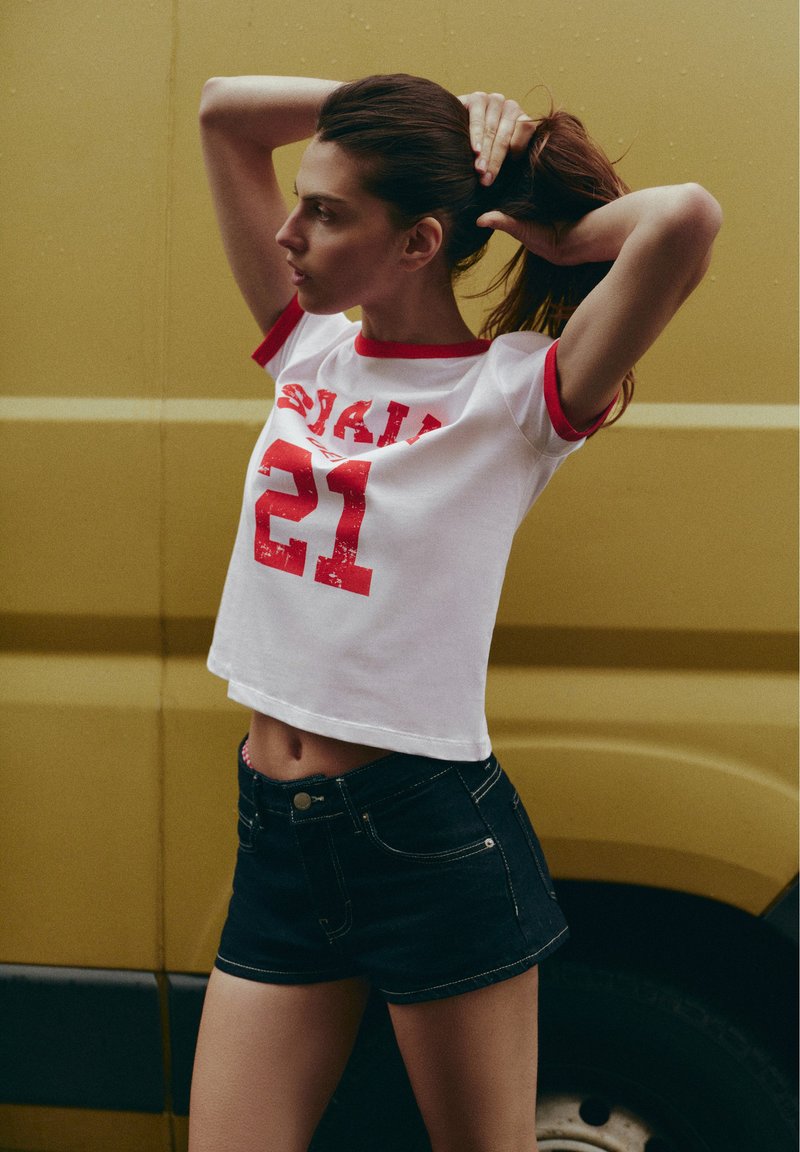 Jeune femme aux cheveux foncés portant un top blanc court et un short noir, attachant ses cheveux, debout devant un véhicule jaune.