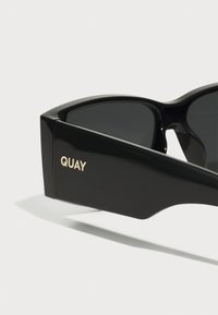 Sorte solbriller med vinkelret design, med en skinnende sort ramme og mørke linser. Guld "QUAY" logo på siden tilføjer en karakteristisk detalje.