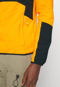 Giacca in pile arancione con accenti neri, tasca con zip, polsini elasticizzati e cuciture a contrasto. Texture morbida, design leggero.