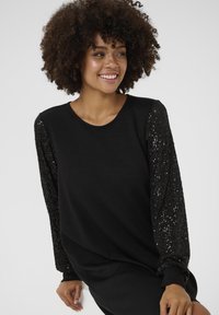 Crna dukserica s dugim, teksturiranim rukavima ukrašenim crnim šljokicama, okruglim izrezom i ležernim krojem. Glatka tkanina na tijelu.