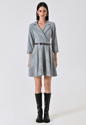 Grau kariertes Kleid mit Reverskragen und Dreiviertelärmeln, tailliert mit einem schwarzen Gürtel. Getragen mit kniehohen schwarzen Stiefeln.