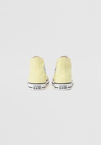 Gelbe High-Top-Sneaker mit glattem Textil-Obermaterial, schwarzer Gummisohle und zweifarbigen schwarzen und weißen Streifenakzenten am unteren Rand. Converse-Logo sichtbar.