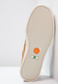 Sko-sula i ljusbrunt gummi med ett texturerat, vågigt mönster. Har en cirkulär orange logotyp och en grön rektangulär knappdesign.