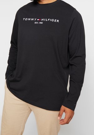 Mann trägt ein langärmliges schwarzes Tommy Hilfiger Shirt mit Logo und beige Hose, steht mit einer Hand in der Tasche.