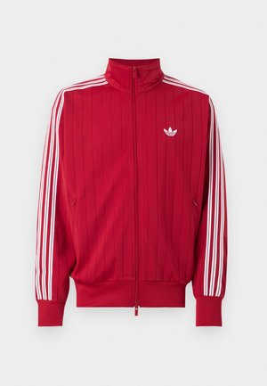 Rød trackjakke med lynlås med hvide Adidas tre striber ned langs ærmerne og lille hvid trefoil-logo på venstre bryst.