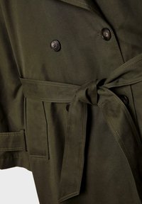 Olivengrøn bælte-trenchcoat med dobbeltknaplukning, sorte knapper og frontlommer. Lavet af en glat, struktureret stof.