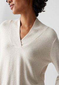 Beige gebreide trui met een V-halslijn en een ontspannen pasvorm. Heeft een subtiele textuur en een zachte stof, die de soepele draping benadrukt.