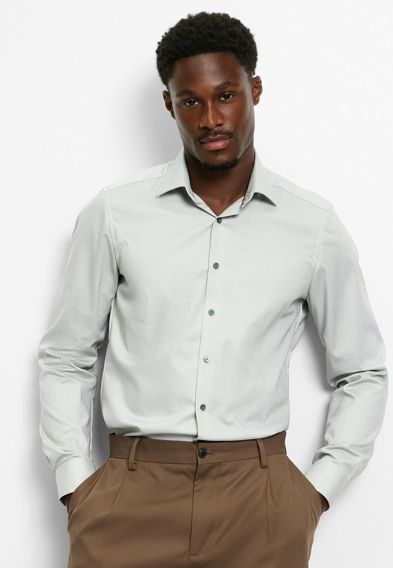 Lichtgrijs langemouwen shirt met een textuurpatroon, knoopsluiting aan de voorkant en kraag, gecombineerd met een bruine broek.