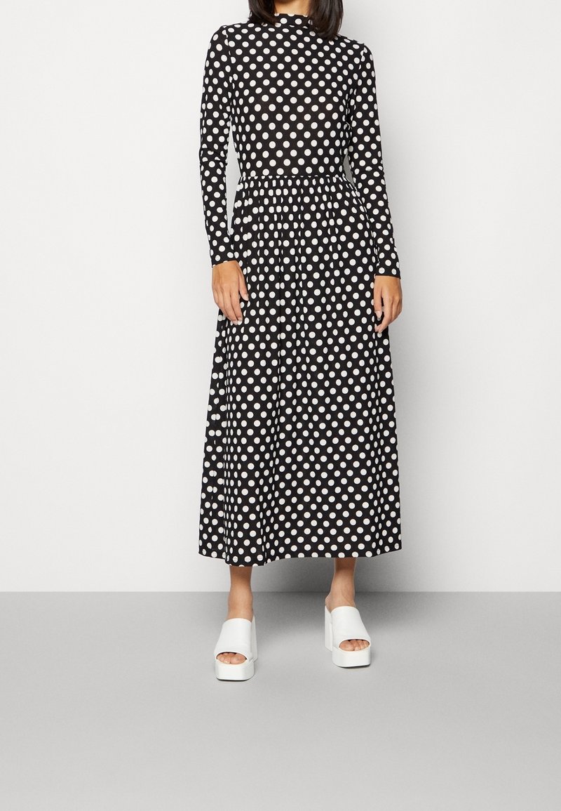 Femme portant une robe midi noire à manches longues avec des pois blancs et des sandales blanches à plateforme à bout ouvert, debout devant un fond uni.