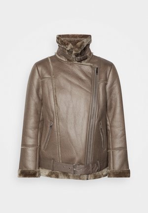 Kvinde iført brun shearlingjakke med bælte, hvid sweater, sorte bukser med vide ben og sorte spidse hælede sko mod ensfarvet baggrund.