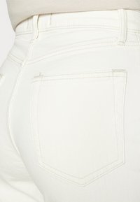 Jeans in denim bianco con dettagli sulla tasca posteriore. Cuciture spesse e una sottile texture visibile. Fit a vita alta con un design pulito e aderente.