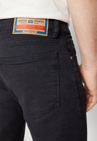Svarta denimjeans med en synlig söm på bakfickan, med en tan-färgad läderetikett som visar text i röd och blå.