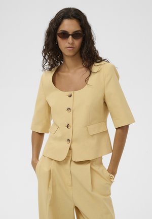 Nő bézs színű, rövid ujjú, gombos blézerben, hozzáillő, magasderekú, pliszírozott nadrágban és sötét napszemüvegben, kezét zsebében tartva.