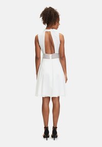 Vit klänning med ärmlös design, öppen rygg, åtsittande midja med intrikata detaljer och en utsvängd kjol. Bärs med svarta högklackade skor.