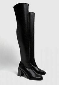 Vysoké černé kožené boty s blokovými podpatky a čtveratými špičkami, stojící vzpřímeně na jednoduchém bílém pozadí.