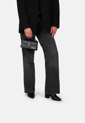 Borsa in pelle nera con un design strutturato e manico superiore, tenuta da una persona che indossa un blazer nero e jeans a zampa d'elefante grigi.