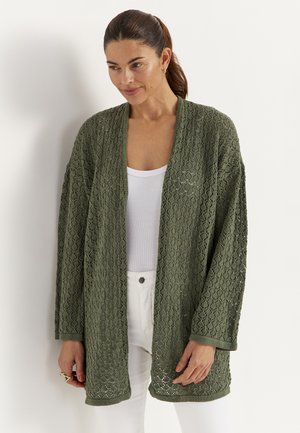 Frau trägt eine olivgrüne, offen geschnittene Strickjacke mit Struktur über einem weißen Tanktop und weißen Hosen, steht vor weißem Hintergrund.