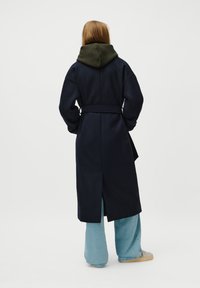 Palton lung de culoare bleumarin cu talie îmbrăcată, glugă de culoare verde închis și spate despicat. Asortat cu blugi largi de culoare bleumarin deschis. Textură moale.