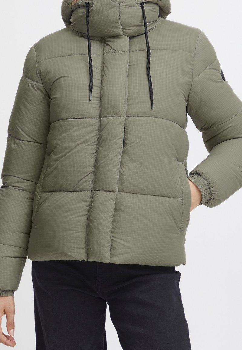 Veste matelassée vert olive avec fermeture éclair, col montant et cordons ajustables. Caractérisée par une texture matelassée et des poches latérales.