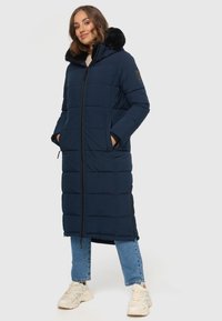 Marineblauer langer Daunenmantel mit einer Kapuze aus Kunstfell, gestepptem Design, Frontreißverschluss und Seitentaschen. Kombiniert mit Jeans und hellen Sneakers.