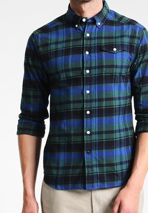 Mężczyzna ubrany w niebiesko-zieloną koszulę w kratę z podwiniętymi rękawami oraz beżowe spodnie, stojący na białym tle.