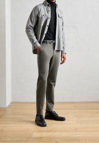 Šedá košile na knoflíky se dvěma náprsními kapsami, černý rolák, olivově zelené slim-fit kalhoty a černé šněrovací boty na dřevěné podlaze.