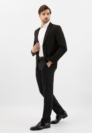 Mann im schwarzen Anzug mit weißem Hemd und schwarzen Lederschuhen steht mit einer Hand in der Tasche und schaut seitlich.
