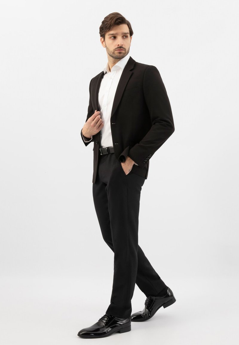 Homme en costume noir avec chemise blanche et chaussures habillées noires, debout avec une main dans la poche et regardant sur le côté.