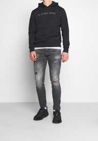 Zwarte pullover hoodie met het "G-STAR RAW" logo, gecombineerd met versleten grijze jeans en zwarte sneakers. Opvallende details zijn een voorkantzak en capuchon.
