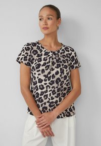 T-shirt à manches courtes en beige clair avec un imprimé léopard noir, présentant un col ras du cou et un léger plissage à l'avant. Le tissu semble lisse.