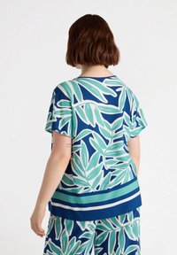 Top a maniche corte con sfondo blu e grandi motivi di foglie verdi e bianche, completato da un orlo a righe. Tessuto leggero.