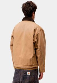 Giacca in tela marrone con colletto in velluto, caratterizzata da una vestibilità ampia e cuciture visibili, indossata sopra pantaloni scuri con un patch del logo.
