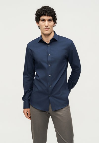 Άνδρας με σγουρά μαύρα μαλλιά που φοράει εφαρμοστό μπλε σκούρο πουκάμισο με κουμπιά και γκρι παντελόνι σε απλό ανοιχτό φόντο.