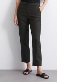 Pantalons taillés verts foncés avec une coupe droite, poches latérales et une texture lisse. Associés à des sandales noires à enfiler.