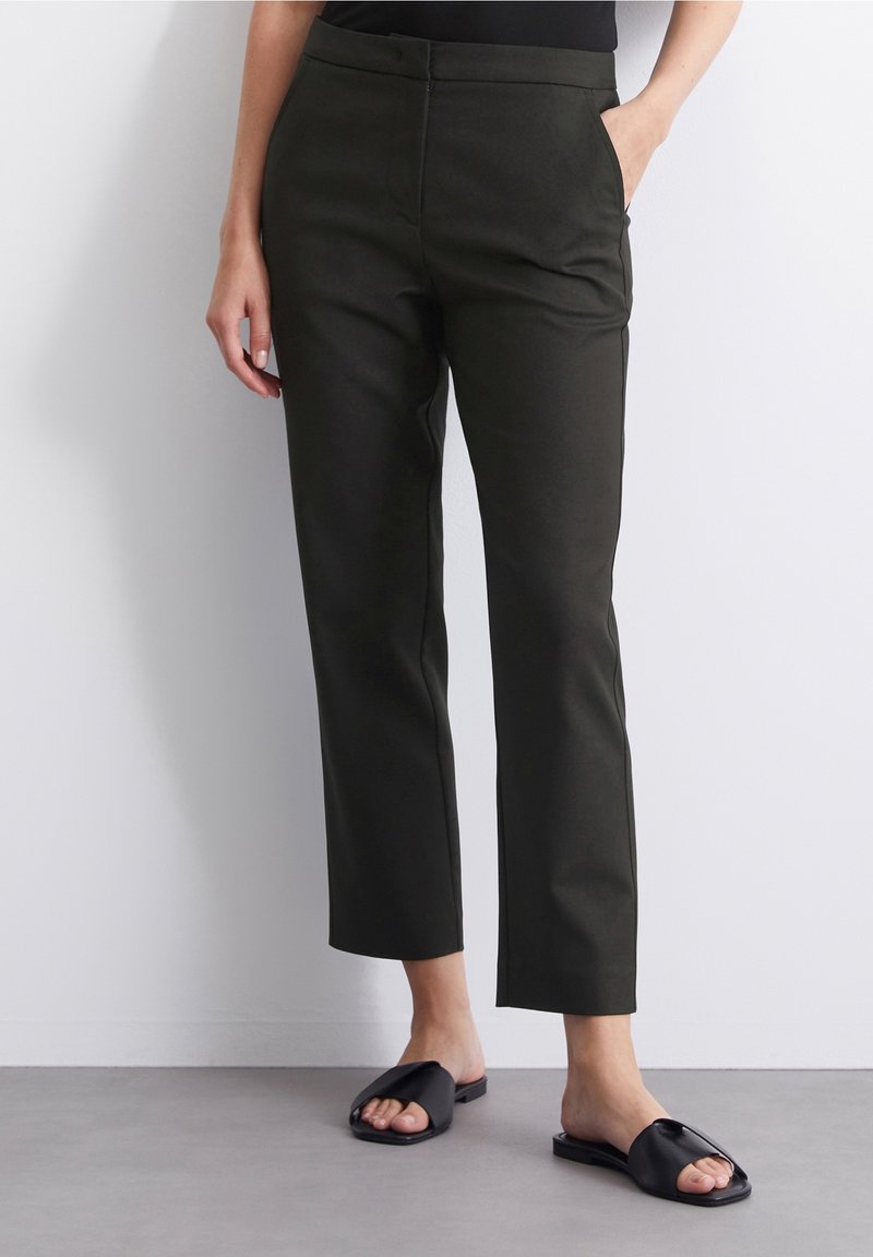 Pantalons taillés verts foncés avec une coupe droite, poches latérales et une texture lisse. Associés à des sandales noires à enfiler.