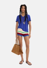 Niebieska koszulka z dekoltem w serek, pasiaste, wielokolorowe szorty z czerwonymi, zielonymi i żółtymi akcentami, brązowa zamszowa torba, czarne klapki, teksturowane włosy.